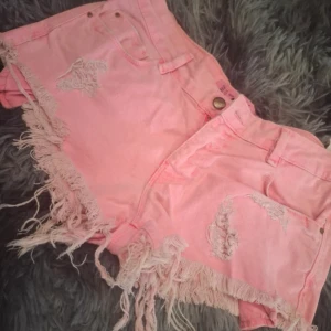 Rosa jeansshorts med fransar - Säljer ett par ljusrosa jeansshorts från Lee med slitna detaljer och långa fransar längs bensluten. Shortsen har klassisk femficksdesign och knappgylf. Perfekta för sommaren!