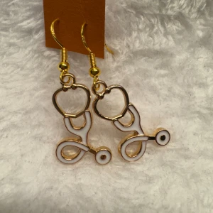 Guldiga örhängen med stetoskopdesign - Unika örhängen i form av små stetoskop med guldfärgade detaljer och vit emalj. Perfekta för dig som gillar medicin eller vill ha ett annorlunda smycke. Hängande modell med krok. Krokarna är i sterling silver 