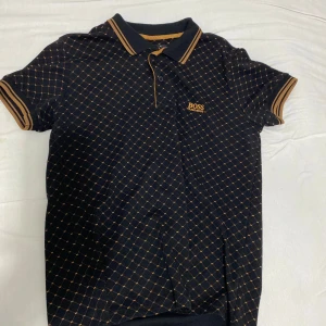 Svart pikétröja med orange rutmönster från Hugo Boss - Säljer en svart pikétröja från Hugo Boss med orange rutmönster och orange detaljer på krage och ärmslut. Klassisk pikékrage och korta ärmar. Boss-logga broderad på bröstet. Perfekt för en stilren och sportig look.