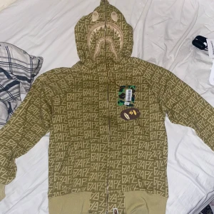 Grön BAPE zip hoodie  - Säljer en grön sällsynt zip hoodie från BAPE med all-over logomönster och hajmotiv på huvan. Jackan har dragkedja hela vägen upp, ribbade muddar och klassisk BAPE-tag vid nederkanten. Perfekt för dig som gillar streetwear och unika detaljer. . storlek M. kom med frågor om storlek och pris!