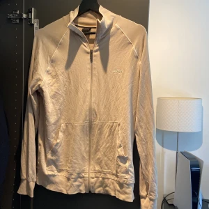 Beige ziptröja från BOSS - Snygg beige ziptröja från BOSS med broderad logga på bröstet. Tröjan har långa ärmar, dragkedja framtill och två fickor. Perfekt för en avslappnad och stilren look.