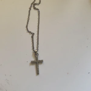 Silverfärgat halsband med korshänge - Säljer ett stilrent halsband i silverfärg med ett korshänge dekorerat med små glittrande stenar. Perfekt accessoar för dig som gillar klassiska smycken med lite extra bling.