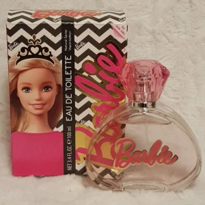Barbie parfym eau de toilette 100ml oanvänd - Säljer en söt parfym från Barbie med en genomskinlig flaska och rosa diamantformad kork. Flaskan har Barbie-loggan i rosa och guld. Förpackningen har svartvitt zickzackmönster, guldiga detaljer och en bild på Barbie med krona och rosa tröja.