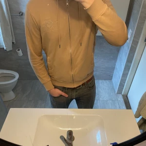 Beige ziphoodie från GANT - Säljer en beige hoodie från GANT med dragkedja och broderad logga på bröstet. Jackan har huva med dragsko och fickor på sidorna. Perfekt för en avslappnad stil.