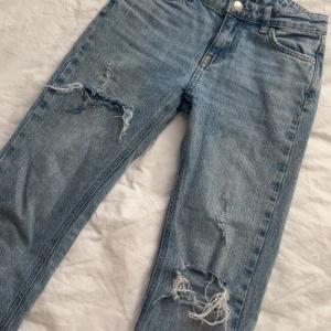 Blå jeans med slitningar från Gina Tricot - Snygga blå jeans från Gina Tricot med coola slitningar på benen. De har en klassisk femficksdesign och knappgylf. Perfekta för en avslappnad stil.