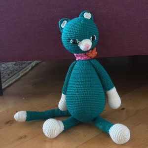Virkad katt i turkos och vitt - Säljer en handgjord virkad katt i turkos med vita tassar, nos och öron. Katten har svarta ögon, rosa nos och bär en rosa och orange virkad blomma runt halsen. Perfekt som present eller dekoration.