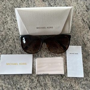 Bruna solglasögon från Michael Kors - Snygga solglasögon från Michael Kors i brun tonad plast med lätt cat eye-form. Märkeslogga på skalmarna och mörka glas. Kommer med originalfodral, putsduk och tillbehör. Perfekt accessoar för soliga dagar.