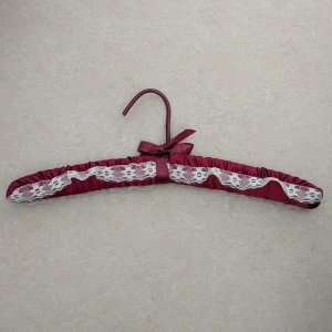 Bordeauxfärgad klädgalge med spetsdetaljer - Säljer en klädgalge i bordeauxfärgat satintyg med dekorativ vit spets längs hela längden. Klädgalgen har en rosett i samma tyg och en mjuk, vadderad känsla. Perfekt för att ge en romantisk touch.