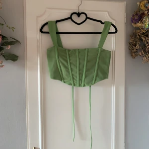Grön korsettopp från Gina Tricot - Snygg grön korsettopp från Gina Tricot med breda axelband och snörning i ryggen. Perfekt för att ge en stilren och trendig look. Toppen är croppad och har en figurnära passform.