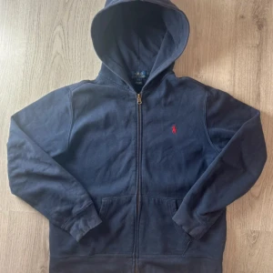 Mörkblå hoodie från Ralph Lauren - Säljer en snygg mörkblå hoodie från Ralph Lauren med dragkedja och huva. Den har en liten röd logga på bröstet och är perfekt för en avslappnad stil. Passar bra till både jeans och joggers.