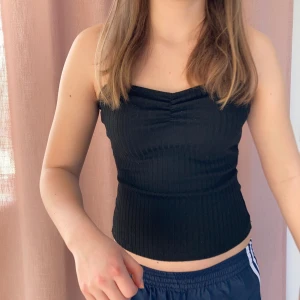 Svart tubtopp - Snygg svart  tubtopp med en smickrande passform. Toppen är axelbandslös och har en lätt rynkad detalj framtill. Perfekt för en stilren look. S-XS