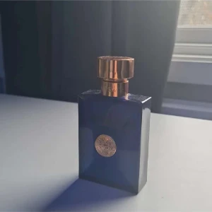 Blå parfymflaska med guldlock - Elegant blå parfymflaska från Versace med ett distinkt guldlock och en dekorativ guldmedaljong på framsidan. Flaskan har en lyxig och stilren design som utstrålar exklusivitet och klass.