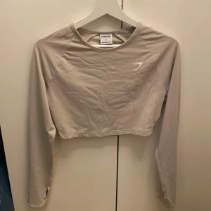 Beige långärmad croppad topp från Gymshark - Säljer en beige långärmad croppad topp från Gymshark. Perfekt för träning med sin bekväma passform och stilrena design. Toppen har en diskret logga på bröstet. Passar utmärkt för dig som vill ha något snyggt och funktionellt att träna i.