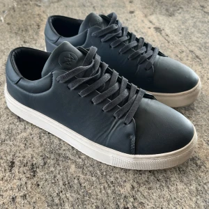 Navy blå sneakers från Kronstadt - Säljer ett par Navy blå sneakers från Kronstadt med vit sula och klassisk snörning. Skorna har en stilren design med rund tå och är tillverkade i syntetmaterial. Perfekta för dig som gillar en enkel och clean look.