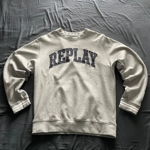 Grå sweatshirt från Replay - Säljer en grå sweatshirt från Replay med tryckt logga på bröstet och patch på ärmen. Tröjan har rund halsringning och ribbade muddar vid ärmslut och nederkant. Perfekt för en avslappnad stil.