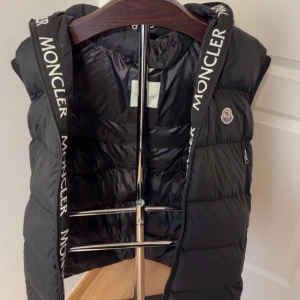 Svart dunväst från Moncler med huva - Snygg svart dunväst från Moncler med avtagbar huva och dragkedja framtill. Västen har två fickor med dragkedja och Moncler-logga på bröstet. Huva och insida har tydlig Moncler-tryck längs kanterna. Perfekt för lager-på-lager-stil.