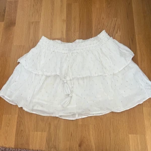 Vit volangkjol med knytband - Söt vit kjol med volang och knytband med tofsar i midjan. Kjolen har en luftig och lätt känsla, perfekt för varma dagar. Den har en kort längd och ett diskret mönster i tyget.