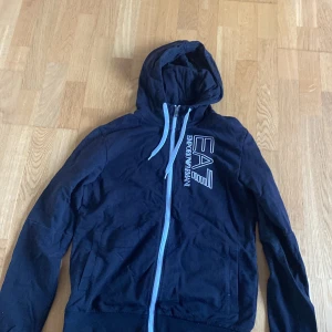 Svart hoodiejacka från EA7 Emporio Armani - Säljer en svart hoodiejacka från EA7 Emporio Armani med dragkedja och huva. Jackan har vita snören i huvan och logga tryckt på bröstet. Perfekt för en sportig och avslappnad stil.