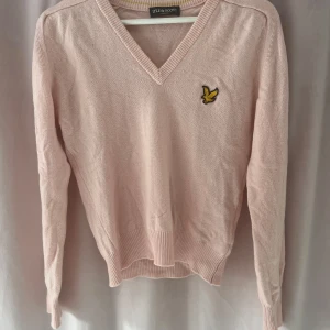 Ljusrosa tröja från Lyle & Scott - Säljer en ljusrosa tröja från Lyle & Scott med v-ringning och ett broderat gult fågelmärke på bröstet. Tröjan har långa ärmar och ribbade muddar vid ärmslut och nederkant. Perfekt för en stilren och avslappnad look.
