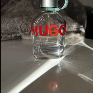 Parfym från Hugo Boss - Säljer en stilren parfym från Hugo Boss med en genomskinlig flaska och röd text. Flaskan har en metallisk kork med en grön rem för enkel användning. Perfekt för den som vill ha en fräsch och modern doft.