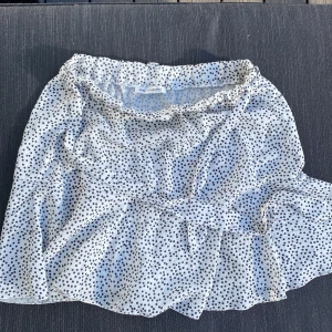 Vit prickig kjol från New Collection - Säljer en söt vit kjol med svarta prickar från New Collection. Kjolen har en volangdetalj och är tillverkad i lätt och luftig polyester. Perfekt för varma dagar! Skönt med skort (shorts/kjol)