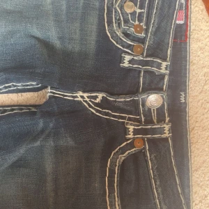 Blå jeans från True Religion - Snygga eftertraktade blå jeans från True Religion med kontrasterande vita sömmar och slitna detaljer. De har en klassisk femficksdesign och knappar med märkets logga. 