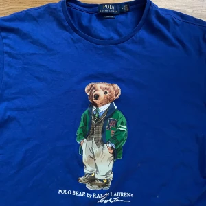 Blå t-shirt med Polo Bear från Ralph Lauren - Säljer en blå t-shirt från Ralph Lauren med en cool Polo Bear-tryck på framsidan. Björnen är klädd i en grön jacka och beige byxor. Perfekt för en avslappnad stil!