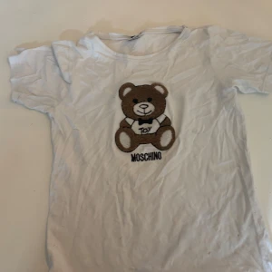 Vit t-shirt med nalle från Moschino - Säljer en söt vit t-shirt från Moschino med en stor broderad nalle på framsidan. Perfekt för en avslappnad stil med en touch av lekfullhet. 🐻