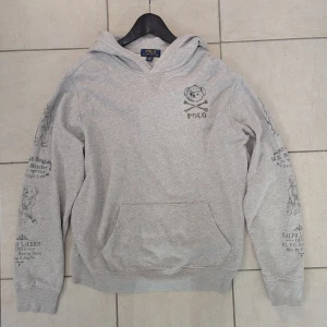 Grå hoodie från Polo Ralph Lauren med björntryck - Säljer en grå hoodie från Polo Ralph Lauren med ikoniskt björntryck på bröstet och tryck på ärmarna. Tröjan har huva, magficka och ribbade muddar. Perfekt för en avslappnad och stilren look. Junior storlek XL motsvarar Small i vuxen storlek