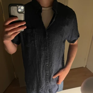 Mörkblå linneskjorta med kort ärm - Snygg mörkblå skjorta i linne med korta ärmar och klassisk krage. Skjortan har knappar framtill och en bröstficka. Perfekt för varma dagar och enkel att matcha med jeans eller shorts.