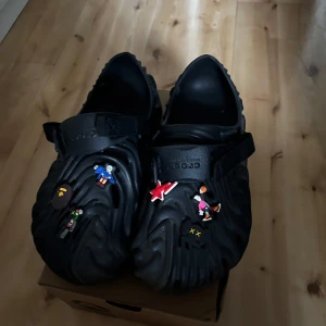 Svarta Crocs Pollex Clog med figurer - Säljer ett par svarta Crocs Pollex Clog med unika figurer och detaljer på ovandelen. Skorna har en vågig design och justerbara remmar för extra komfort. Perfekta för dig som vill sticka ut med dina skor!