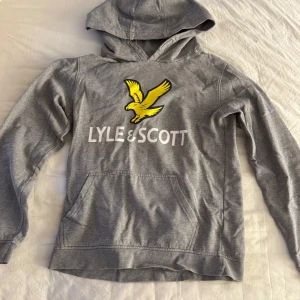 Grå hoodie från Lyle & Scott - Säljer en grå hoodie från Lyle & Scott med en gul logga och text framtill. Tröjan har huva och en stor magficka. 
