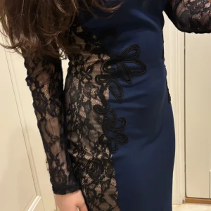 Marinblå klänning med spetsdetaljer - Säljer en marinblå klänning med svarta spetsärmar och spets längs ena sidan. Klänningen är tight och har långärmade spetsärmar som ger en elegant look. Perfekt för dig som vill sticka ut med snygga detaljer.