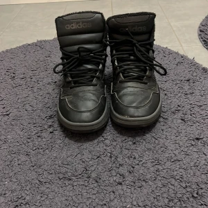 Svarta Adidas boots med snörning - Säljer ett par svarta Adidas boots med högt skaft och snörning. Skorna har klassiska Adidas-ränder på sidorna och är fodrade för extra värme. Perfekta för kyligare dagar och har en sportig look. Jag ska fräscha upp skorna ännu mer och den är i helt ok skick. Storlek 40 2/3