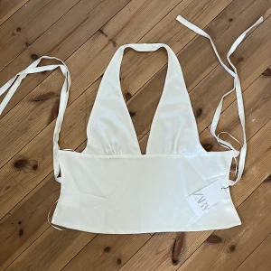 Vit halterneck topp från Zara - Snygg vit halterneck topp från Zara med knytband i ryggen. Perfekt för sommardagar eller en utekväll. Tillverkad i ett mjukt och glansigt material som ger en lyxig känsla. Har en liten fläck.