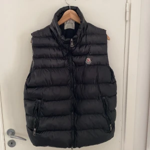 Svart dunväst från Moncler - Säljer en svart dunväst från Moncler med hög krage och två dragkedjefickor framtill. Västen har quiltad design och klassisk Moncler-logga på bröstet. Perfekt att slänga över en hoodie för en clean och sportig look.