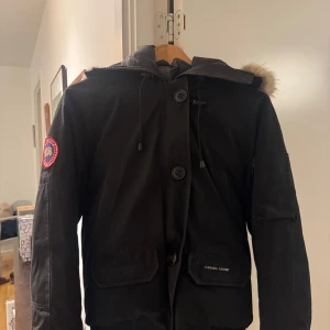 Svart Canada Goose dunjacka med päls - Säljer en svart dunjacka från Canada Goose med ikonisk patch på ärmen och huva med äkta päls. Jackan har stora fickor, dragkedja och knappar framtill samt snörning i huvan. Perfekt för kalla vinterdagar och riktigt snygg streetstil. Kom DM för mer bilder ifall de behövs, har ej kvitto kvar då den köptes för flera år sedan som en present.