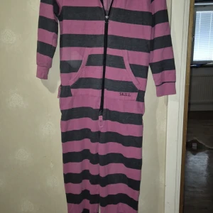 Randig onesie med huva från Skill - Supermysig randig onesie från Skill i rosa och svart. Har huva, långa ärmar, två fickor framtill och dragkedja hela vägen ner. Perfekt för chill dagar hemma eller när du vill vara extra bekväm.