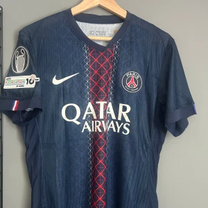 PSG matchtröja D. Doué #14 Nike - Paris Saint-Germain fotbollströja med D. Doué och nummer 14 på ryggen. Mörkblå med röd och vit detalj i mitten, PSG-emblem och Qatar Airways-tryck på bröstet. Korta ärmar, officiella UEFA Foundation-märken på ärmarna och klassisk V-ringning. Helt ny med tagg