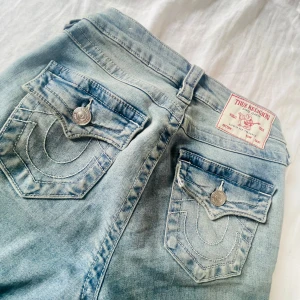 True Religion Becca Bootcut Jeans - Snygga ljusblå bootcut jeans från True Religion, modell Becca med midwaist. Klassiska stora bakfickor med vita kontrastsömmar och silverfärgade knappar. Ikonisk True Religion patch bak. Perfekta för dig som gillar en avslappnad men trendig look.