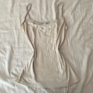 Beige spetslinne från Vero Moda - Supersött beige linne från Vero Moda med tunna axelband och vacker spetsdetalj längs urringningen. Perfekt att styla lager på lager eller som det är. Mjukt material och feminin känsla.