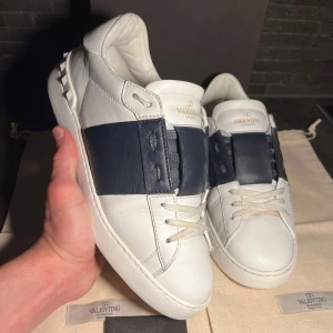 Valentino Garavani Open sneakers vit/marin - Snygga Valentino Garavani Open sneakers i vitt skinn med bred marinblå läderdetalj över snörningen. Klassisk rund tå, platt sula och diskreta nitar bak på hälen. Lyxig känsla och stilren design, perfekta för dig som gillar exklusiva sneakers.