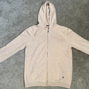 Beige hoodie från Tresanti med dragkedja - Säljer en stilren beige hoodie från Tresanti i mjuk bomullsmix. Tröjan har huva med snörning, hel dragkedja framtill och två fickor. Perfekt för chill dagar och enkel att matcha med jeans eller joggers. Långärmad och med diskret logga vid fickan.