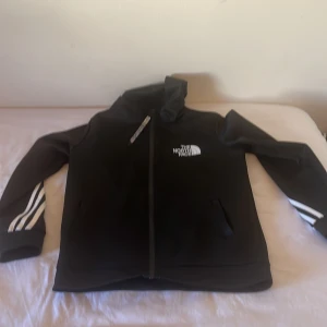 Svart vindjacka från The North Face - Svart vindjacka från The North Face med vit logga på bröstet och vita ränder på ärmarna. Jackan har dragkedja framtill, huva och två fickor. Perfekt för dig som gillar sportig och clean stil.