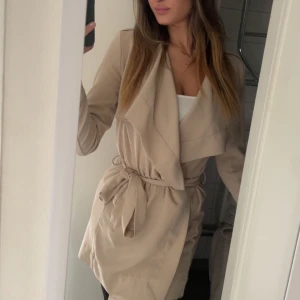 Beige kappa från Object, S - Snygg beige kappa från Object med skärp i midjan. Jackan har långa ärmar, knappar på axlarna och en avslappnad passform och slutar över knäna. Är i mycket väl skick.  Perfekt för vår och höst när du vill ha en stilren look.