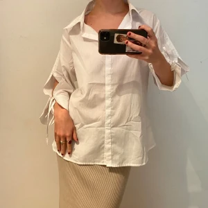 Vit skjorta med snörning på ärmar - Stilren vit skjorta i bomull med klassisk krage och unika snörningsdetaljer på ärmarna. Skjortan har en loose passform och långa ärmar som kan justeras med knytband. Perfekt för dig som gillar en avslappnad men trendig look.