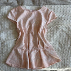 Ljusrosa blus med volangkant - Supersöt ljusrosa blus med diskret mönster och volangkant nertill. Blusen har rund halsringning, korta ärmar och figurnära passform upptill som släpper ut i en peplum-liknande volang. Perfekt för dig som gillar romantisk och feminin stil. Jättefin till jeans eller kjol på sommaren. Men har väl blivit för stark för den passar inte runt mina biccar längre tyvärr.