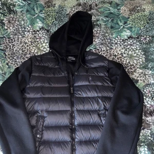 Polo Ralph lauren cardigan - Säljer en svart cardigan från Polo Ralph Lauren med huva och dragkedja. Cardiganen har quiltad front i glansigt material, släta svarta ärmar och broderad logga på bröstet. Perfekt för höst och vinter, riktigt clean och stilren look. Jackan är i bra skick med inga defekter vad jag kan se. Jackan säljs inte längre och är väldigt sällsynt därför krävs ett rätt högt pris.