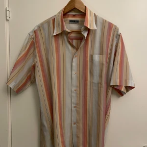 Randig kortärmad skjorta Basic Line - Snygg kortärmad skjorta från Basic Line i storlek L. Skjortan har ett färgglatt randigt mönster i beige, rött, orange och grått. Klassisk krage, bröstficka och knappar framtill. Perfekt för dig som gillar retrovibbar och vill sticka ut lite extra.