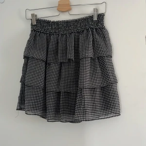 Svartvit rutig volangkjol H&M XS - Supersöt kort kjol från H&M i storlek XS med svartvitt rutigt mönster. Kjolen har flera volanger och en smockad midja som gör den bekväm och lätt att bära. Tillverkad i mjuk polyester som ger ett luftigt och flowy fall.